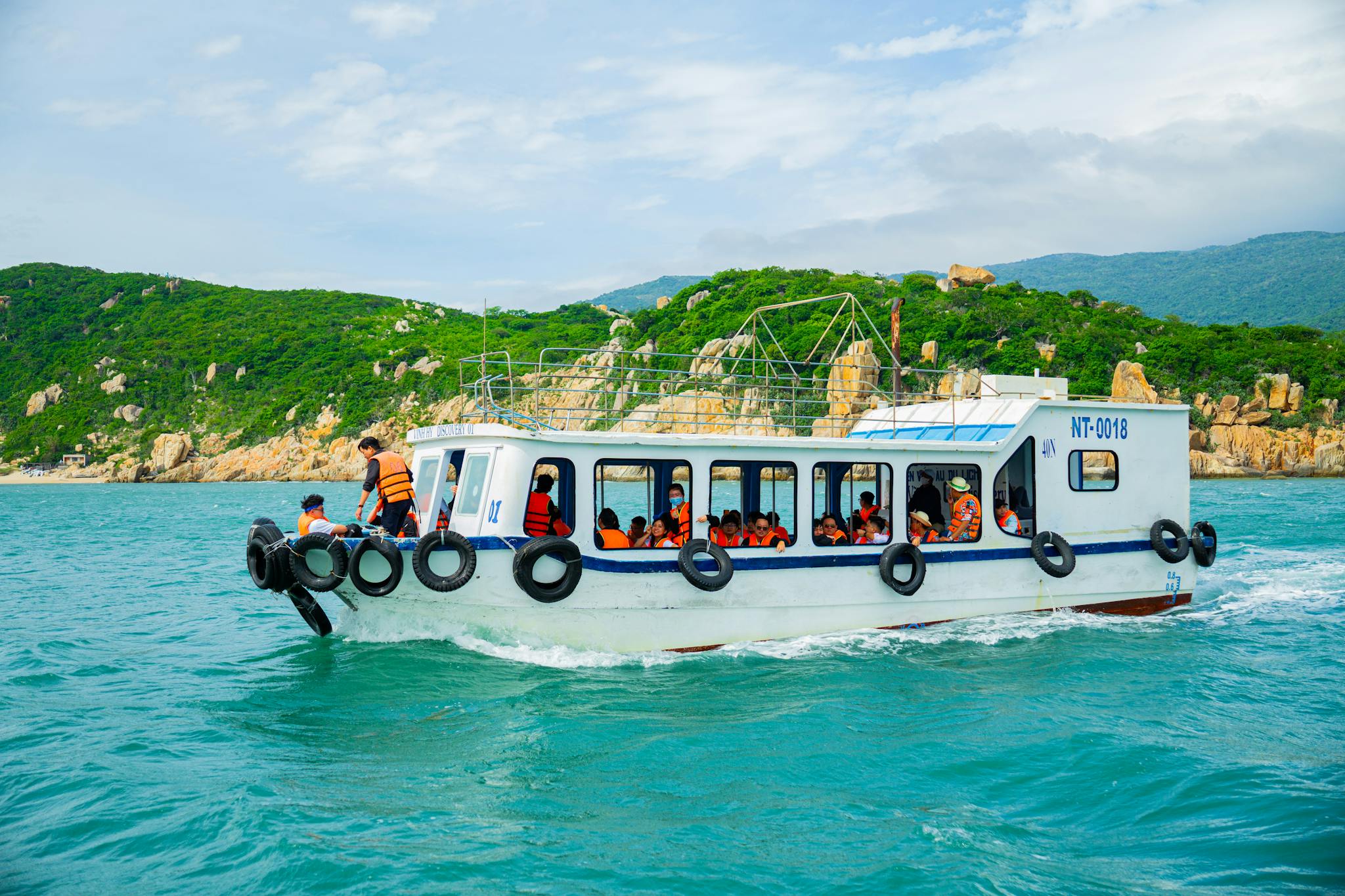 A boat filled with tourists in life jackets traverses vibrant turquoise waters along a lush, green coastline.